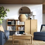Modern living room with wooden bookshelf, blue couch, round mirror, wooden lamp, coffee table, grey curtains.