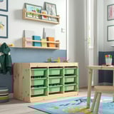 Kids room with TROFAST storage unit. Wooden frame, green plastic boxes. Art, books, and toys displayed. Large, prominent unit with sanded finish.
