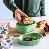 A person puts a sandwich in a 2-tier green lunch box on a table with snacks and grapes, next to a patterned cloth.