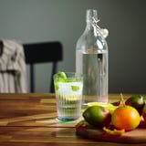 A clear glass filled with water and a mint leaf sits on a wooden table, flanked by limes and oranges. A glass bottle of water is also present.