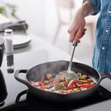 Person cooks colourful stir-fry in black wok with spatula, steam rising, on modern stove.