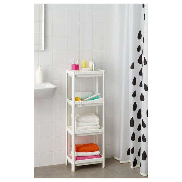 A white, 3-tiered bathroom shelf, VESKEN, holds folded towels and toiletries. Positioned near a sink and shower curtain with black droplets.