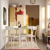 Modern dining area with white table, red lamp, wooden chairs, veggies, bright room, bookshelf.