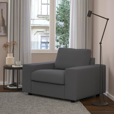 Modern grey chair beside window; tall black lamp, small black side table with dried flowers, mug, and headphones.