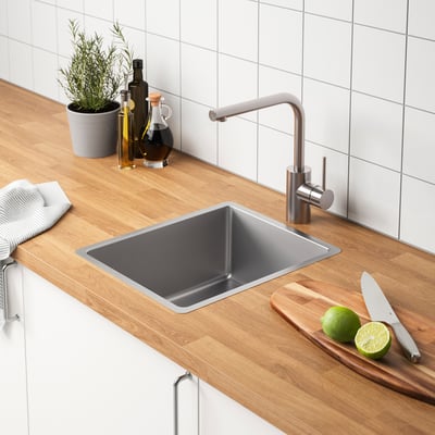 Kitchen counter with stainless steel sink, under-counter tap, wooden cutting board with limes and knife, oil bottles, and potted plant.