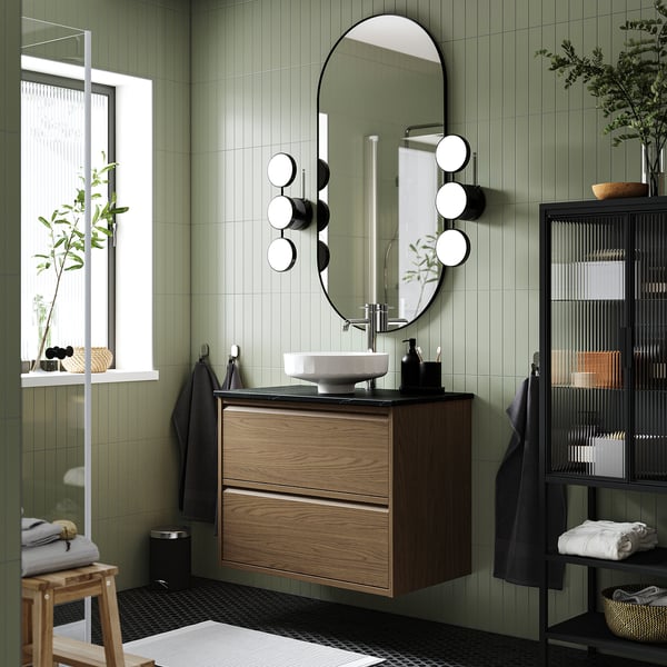 Modern green bathroom with dark wood sink, round basin, mirror with lights. Shelf with towels and plants; small stool.