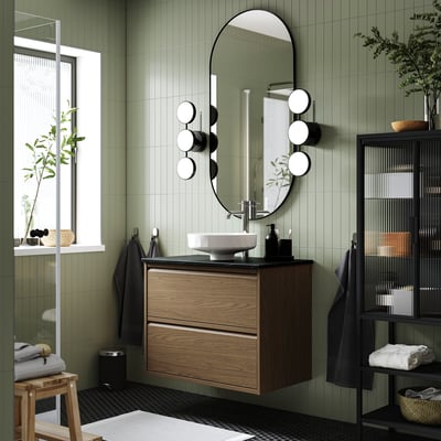 Modern green bathroom with dark wood sink, round basin, mirror with lights. Shelf with towels and plants; small stool.