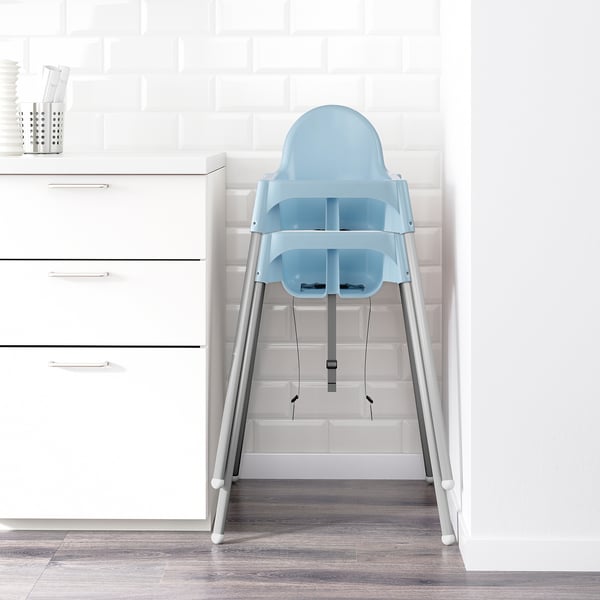 A light blue high chair with a safety belt, designed for children ages 6-36 months, positioned in a white room corner.