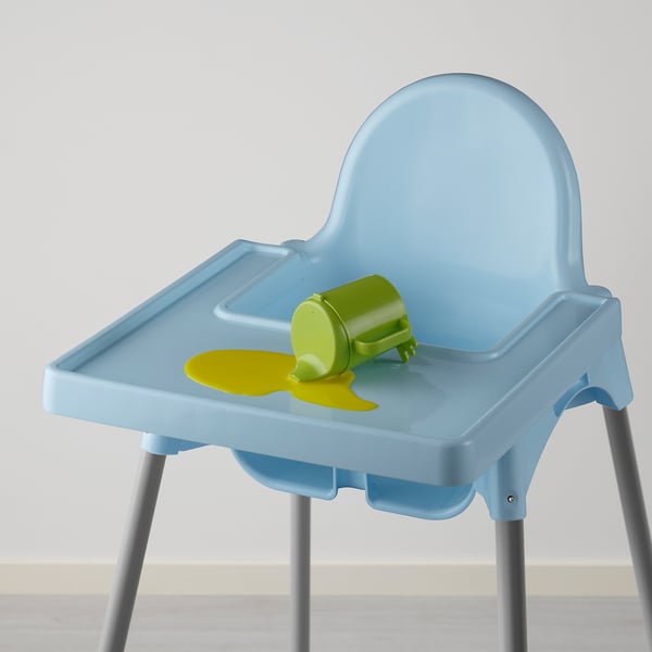 Blue childrens high chair with light grey legs. Green sippy cup lies on tray with yellow spill.