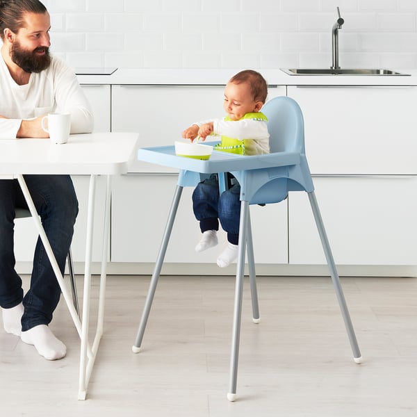 Bearded person watches baby in blue high chair eating from green bowl.