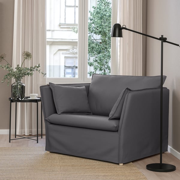 Cosy grey BACKSÄLEN armchair with cushions. Modern, minimalist design. Oversized for comfort. Next to tall window & small black side table with plant.