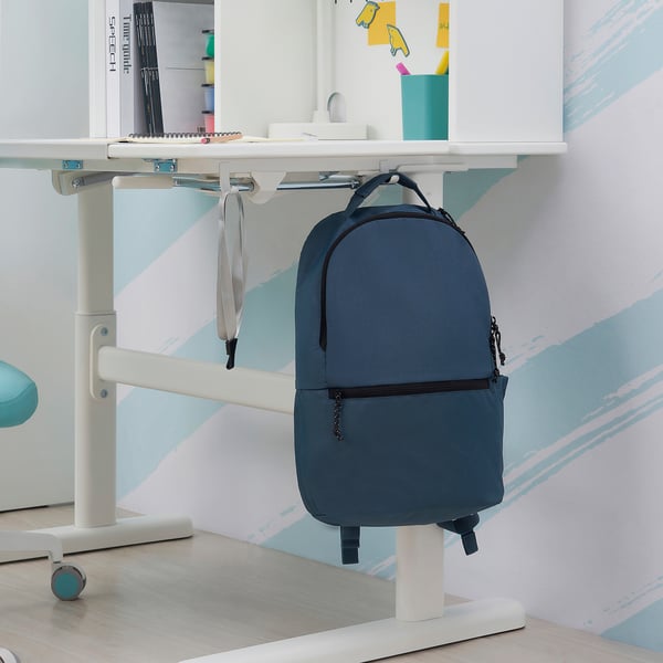 A blue backpack hangs on a white desk with adjustable height and tiltable surface.