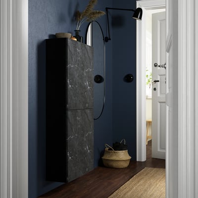 Modern entryway with black cabinet, marble finish, metal legs, and knobs. Its tall, sleek, next to plants and a basket. Wall light illuminates.