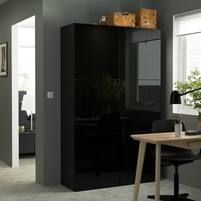Black BESTÅ storage unit, tall and narrow, fits neatly in corner, with woven baskets on top, a plant on the shelf, and a sleek desk beside it.