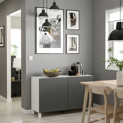 Modern living room with grey BESTÅ storage unit, white top, featuring black pendant lights, framed photos, and wooden chairs.