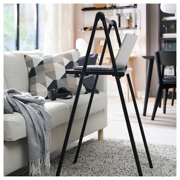 Black adjustable stand-up desk with laptop in living space, next to a grey sofa with a geometric pillow and blanket, offering a comfortable workspace.