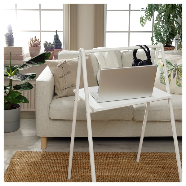 White BJÖRKÅSEN foldable desk, holding a laptop and headphones, placed in front of a beige couch with pillows in a living area with plants.