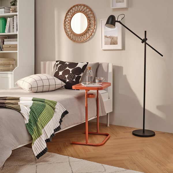 Bedside scene: BJÖRKÅSEN orange laptop stand, black floor lamp, white bed with pillows, green blanket, wooden floor, wall mirror, shelves with books.