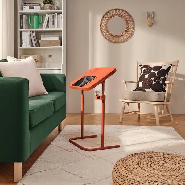 Bright, orange BJÖRKÅSEN laptop stand in cosy room. Its sleek, adjustable design perfectly fits the space between green couch and wooden chair.