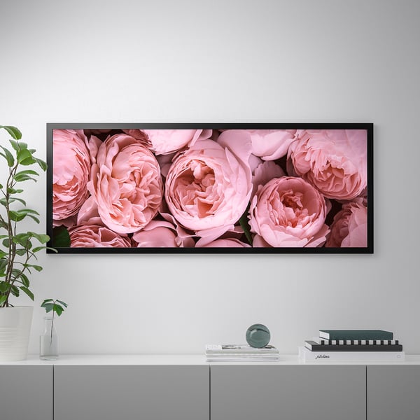 A framed canvas print of pink roses hangs on a wall above a cabinet with decorative items, including a vase with plant and stacked books.