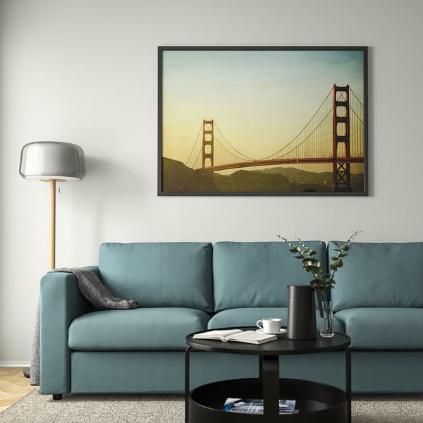 A living room with a large BJÖRKSTA black picture frame showcasing a bridge, a teal couch, round coffee table, plant vase, and lamp.
