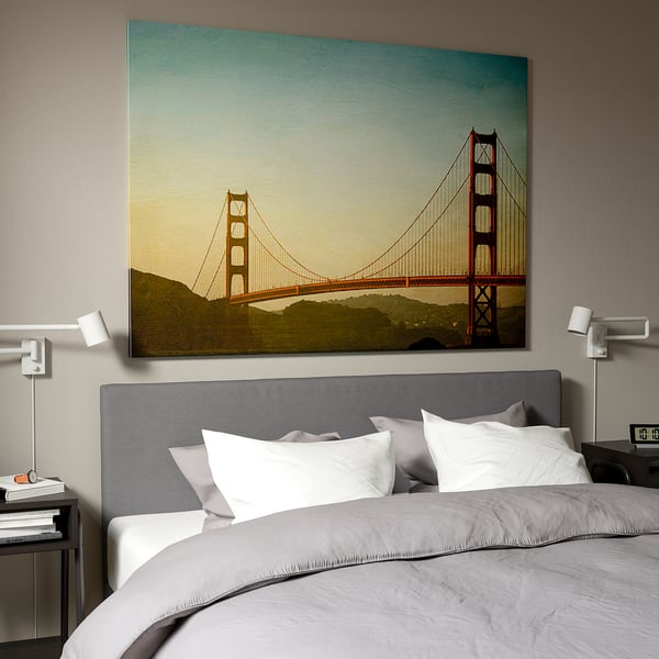 Large bedroom with BJÖRKSTA picture above bed showing golden gate bridge. Grey headboard and white pillows. Modern, minimalist design.