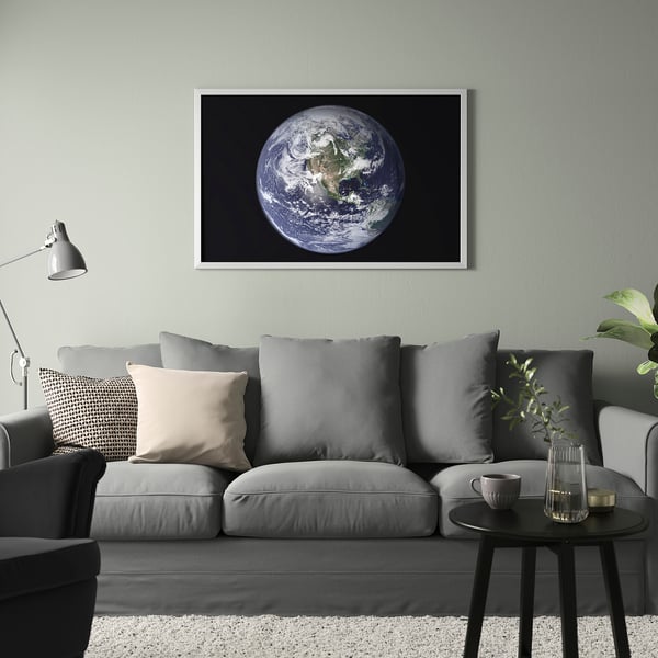 Large framed earth poster above grey sofa. Modern, minimalist living room decor. Grey cushions and black side table. Light grey wall. Vase with plant.