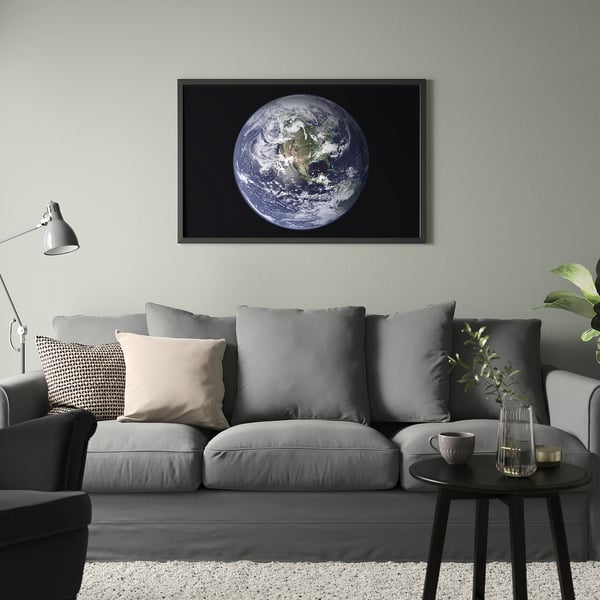 Sunlit earth print in black frame over modern grey couch with neutral pillows and a plant, mug on side table.