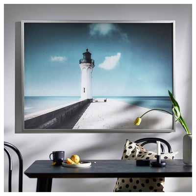 Large wall art with white lighthouse on pier, blue sky. Framed in silver, modern minimalist design.