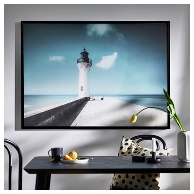 Large framed picture of lighthouse by sea, displayed indoors. Beside table with fruit, cup, and polka dot items.