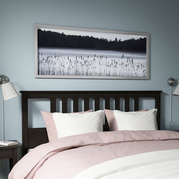 Large BJÖRKSTA framed nature print above cosy bed, grayish-blue wall. Modern, minimalist bedroom decor.
