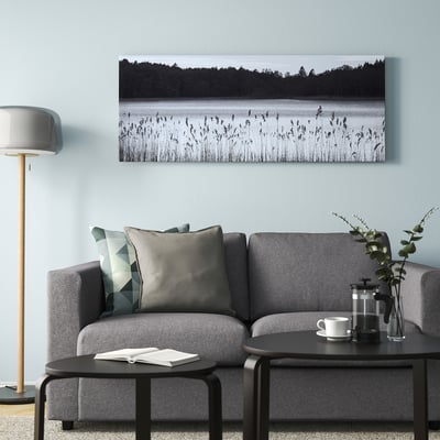 Minimalist living room with grey bjorksta artwork above sofa, calm lake scene, coffee maker, greenery, grey lamp.