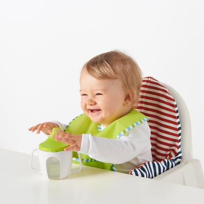 Baby sits in high chair, holding green-handled beaker with white spout, wearing striped outfit and green bib.