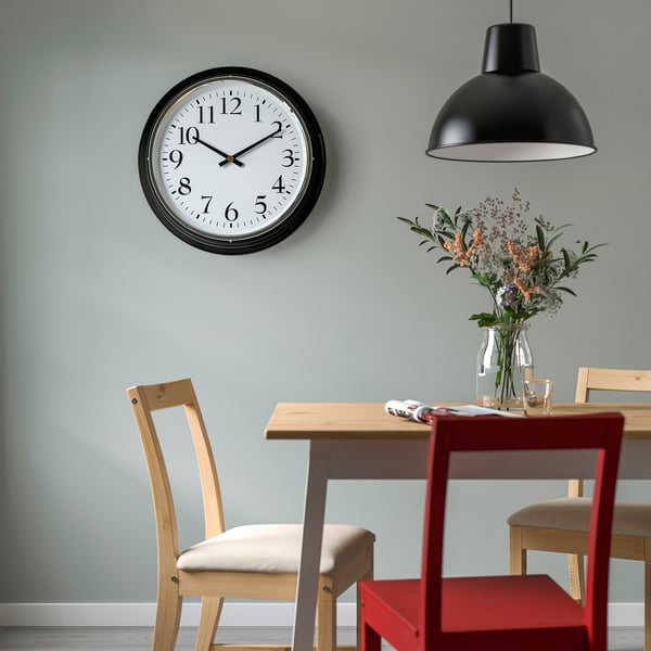 Black BRAVUR wall clock above dining table with flowers and chairs.