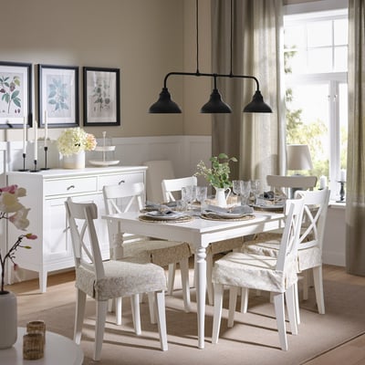 Image shows a dining area with a white table, six matching chairs with beige floral cover, beige curtains, and hanging lights.