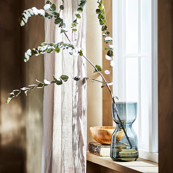 Modern living room with wooden shelf, blue hourglass vase of eucalyptus, books, bowl, and sheer curtains.