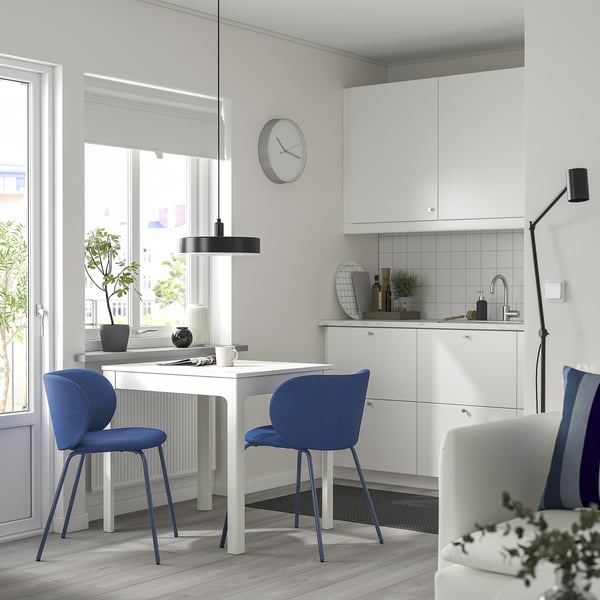 Modern white dining set with blue chairs, sleek design, clock above, plant, kitchen sink, large window, minimalist decor.