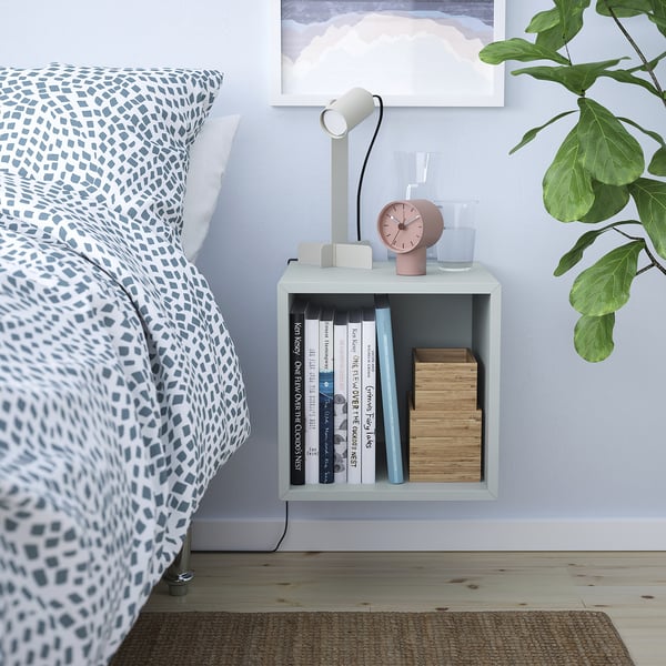 Bedside, simple white EKET cabinet holds books, alarm, and glass. Mounted beside bed, fits well in small space.
