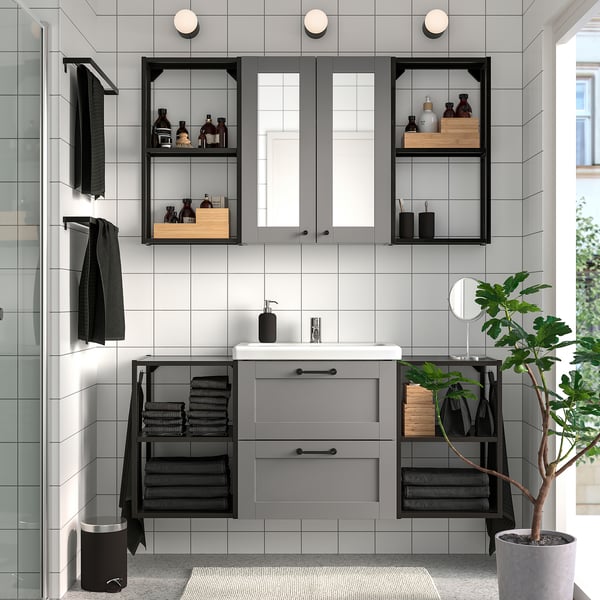 Modern bathroom with grey ENHET vanity, sink, and mirror cabinet. Black accents and open shelving for towels and toiletries. Plant adds greenery.