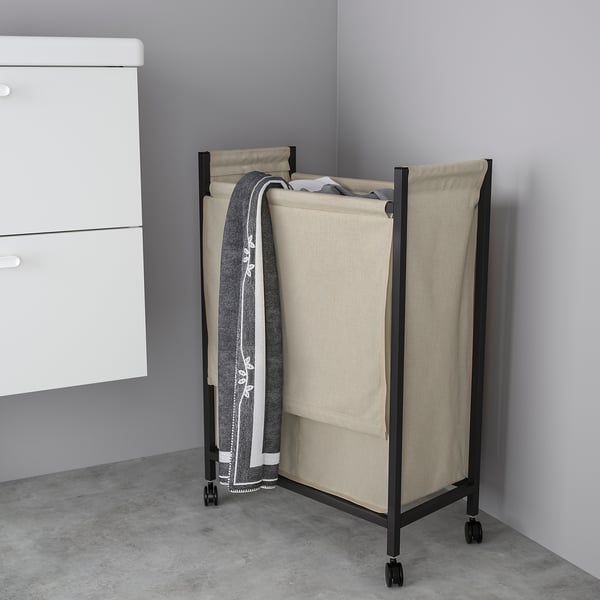 Black ENHET laundry bag on wheels, cornered next to white drawers, holding grey towels.