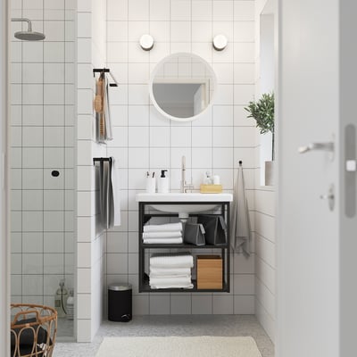 Modern white bathroom: round mirror, black ENHET shelf, ceramic basin, glass shower, hooks, and grey towels.