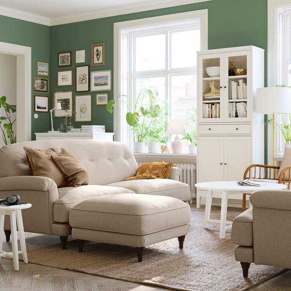 Modern living room with green walls, beige sofa, footstool, wooden chair, potted plants, and framed artwork on windowsill.