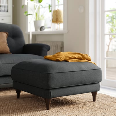 Cosy living room scene with blue upholstered footstool, golden throw, and potted plants.