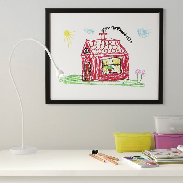 Black FISKBO frame holds red house drawing with green lawn, pink flowers, and yellow sun on white desk with lamp, watercolours, and containers.