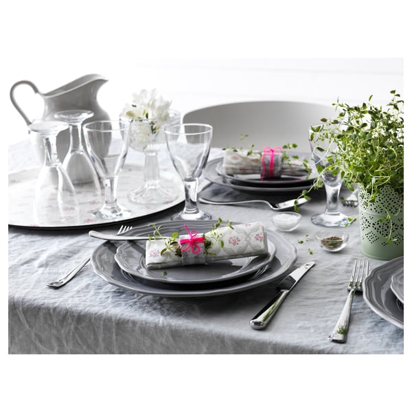 Elegant dining setup with grey plates, silver cutlery, glassware, and decorative wrapped napkins. White flowers in a clear vase as centrepiece.