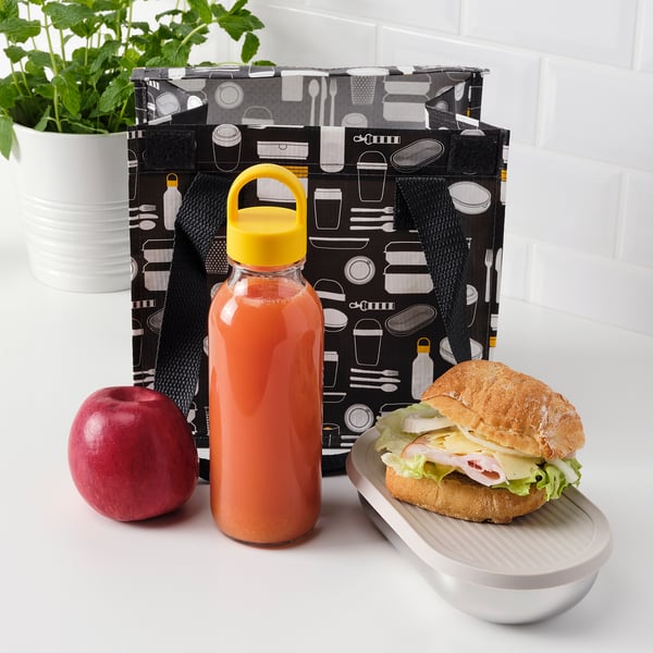 Black bag with utensil patterns holds sandwich, apple, and orange drink in glass bottle, next to a potted plant.