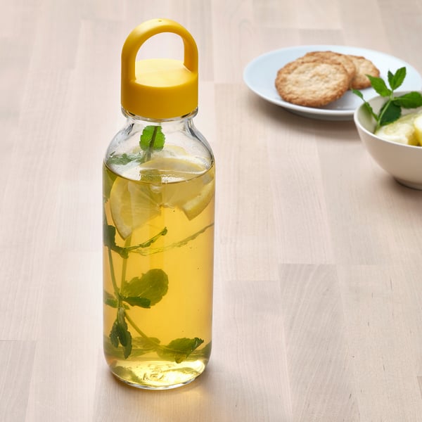 A clear glass bottle with a yellow lid and handle holds lemon-infused water, placed on a wooden table.