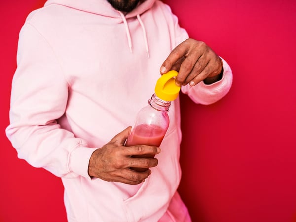 Person in white hoodie holding a pink glass bottle with a yellow lid, pouring a pink drink.