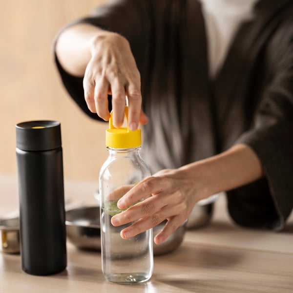 Person holding a yellow-handled, transparent glass bottle on a table with a black bottle nearby.