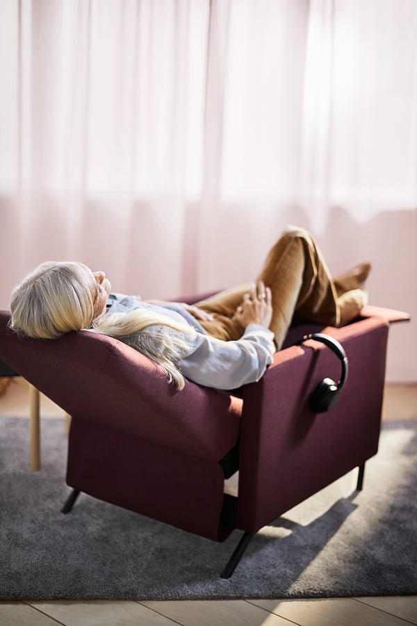 Older person reclining in GISTAD armchair, maroon, listening to headphones.